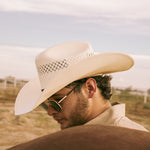 Bearded man wearing a 10X Cowboy Weston black and ivory cowboy hat with a white collared shirt standing behind a brown horse