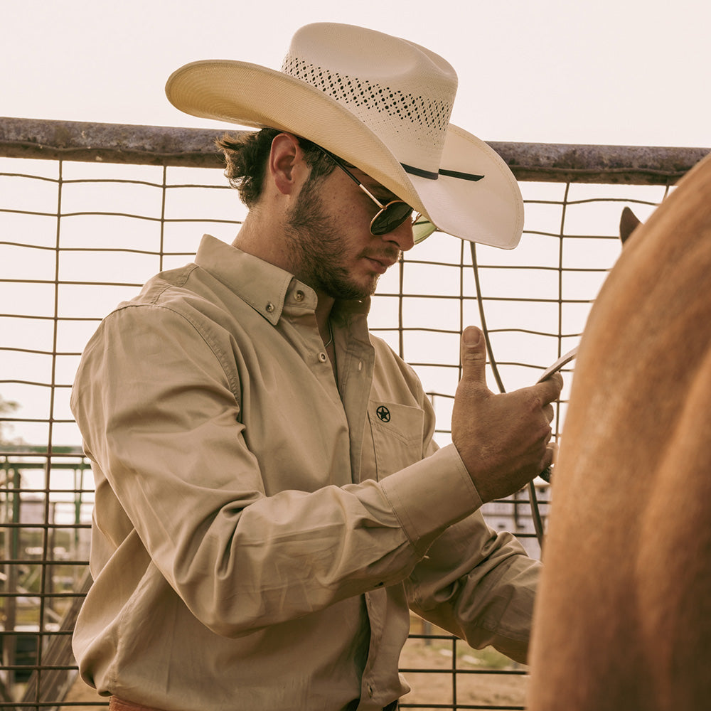 Bearded man wearing a 10X Cowboy Weston black and ivory cowboy hat with a white collared shirt putting saddle on a brown horse