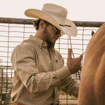 Bearded man wearing a 10X Cowboy Weston black and ivory cowboy hat with a white collared shirt putting saddle on a brown horse