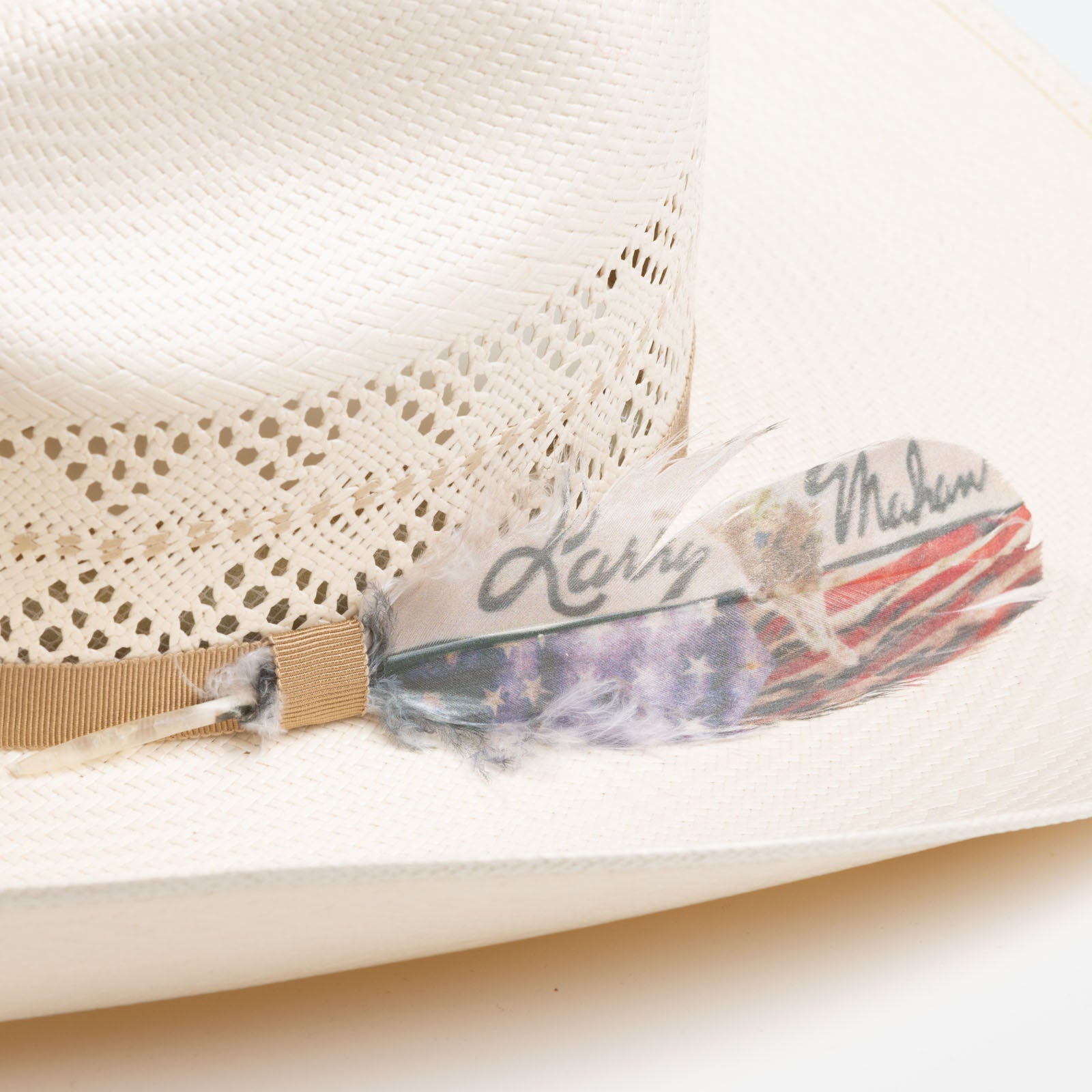10X Tulsa closeup of a light brown band with a gold and black Larry Mahan pin and writing on a feather with a colored American flag along the bottom of the feather