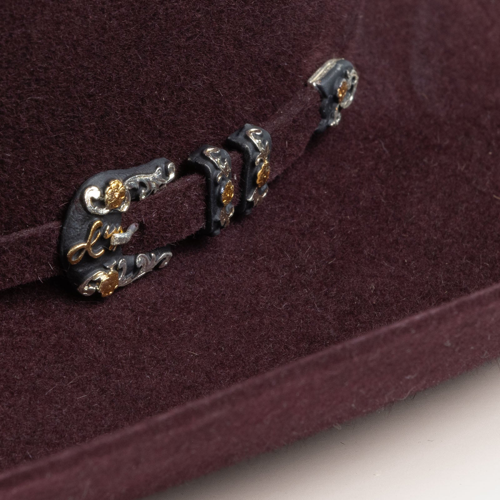 10X Jerarca black cherry cowboy hat closeup of buckle in black with silver and gold floral design with the letter L on a black cherry band