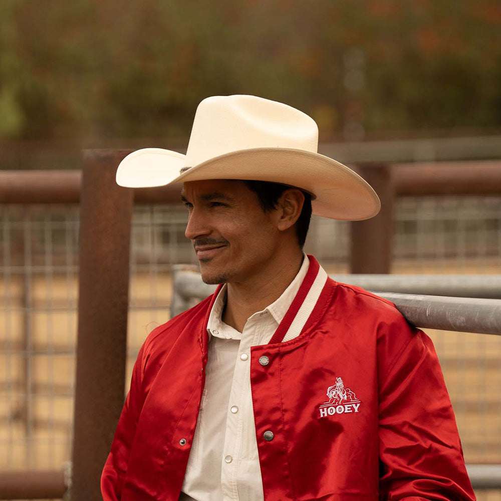 Smiling man wearing a 10X Brindle brown cowboy hat with a bright red jacket and white under shirt standing near metal fences