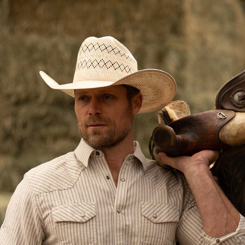 Bearded man wearing a 10X Cowboy Santos tan and ivory cowboy hat with plaid white shirt holding a brown horse saddle over his left shoulder