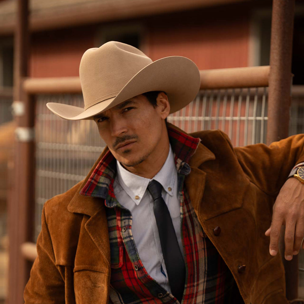Man wearing a 6X El Oro fawn cowboy hat with brown jacket, red and blue plaid long sleeve shirt, white under shirt, and black tie as he leans against a metal fence