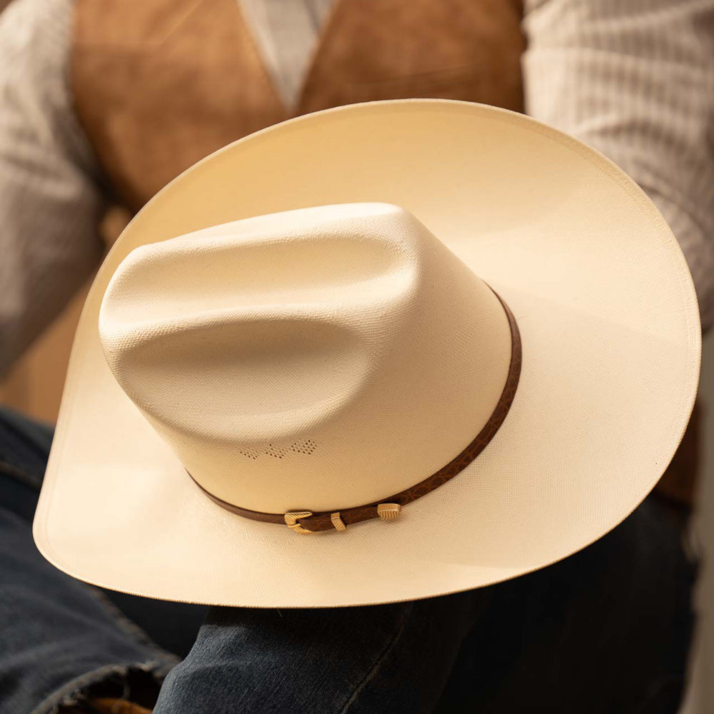 15X El Primero cognac cowboy hat resting in a person's lap with brown band and gold buckle showing the full scope of the top of the hat