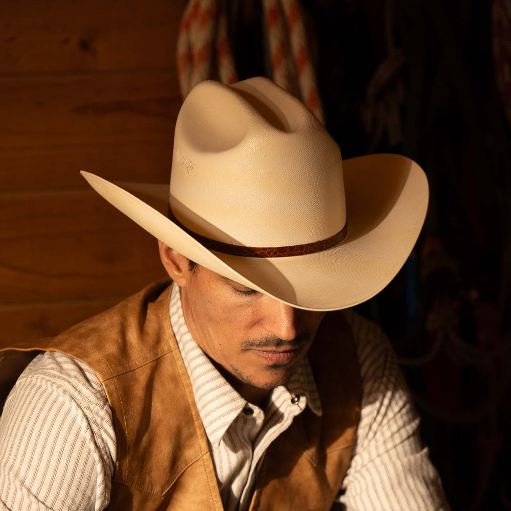 Man wearing a 15X El Primero cognac cowboy hat with brown band, vest, and white striped shirt sitting near a wooden wall