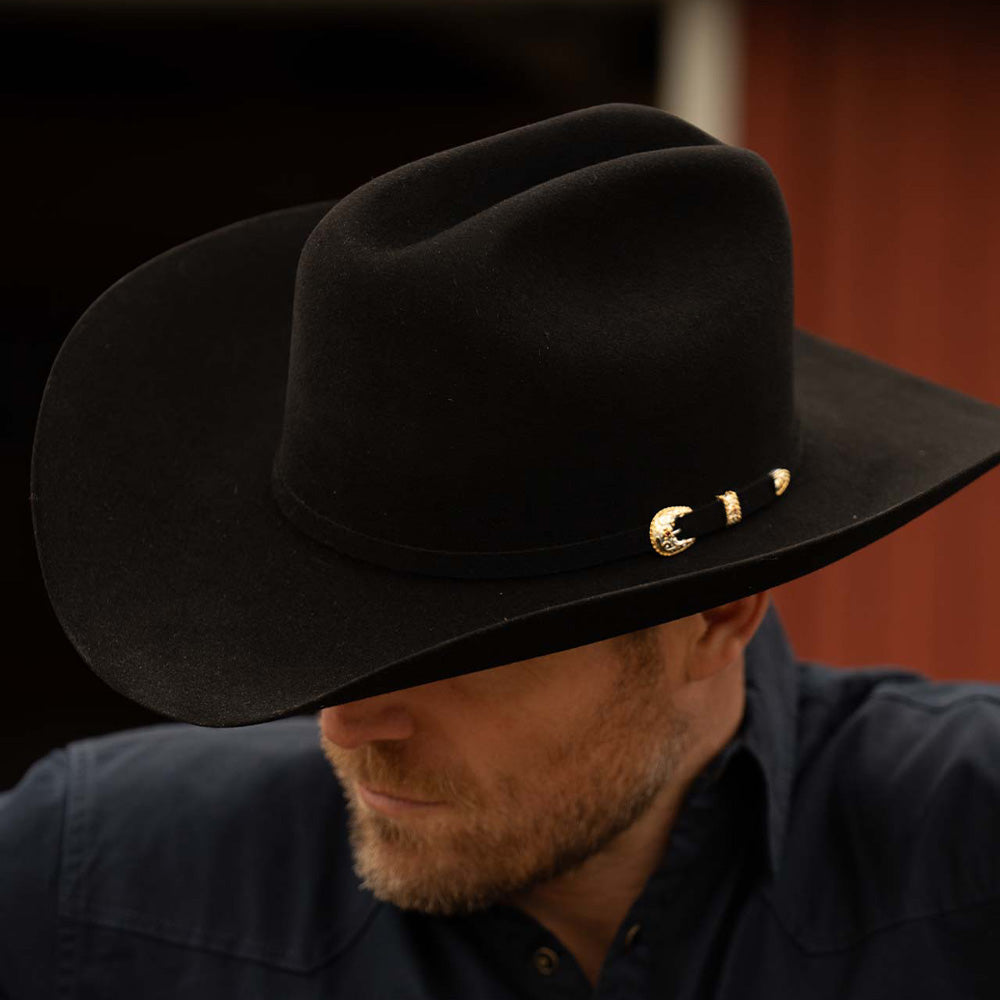 Bearded man wearing a 30X El Rey black cowboy hat and slate-colored long sleeve shirt with his head down and to the left to show the gold buckle on the brown band