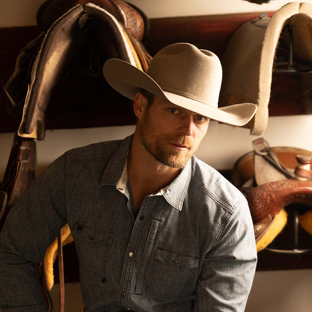 Bearded man wearing a 5X Fort Worth natural cowboy hat and blue long sleeve shirt with horse saddles stacked behind him on a wall