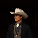 Man wearing a 100X Independencia belly cowboy hat with black jacket, grey shirt and vest, and silver bolo tie with gold horse riding emblems