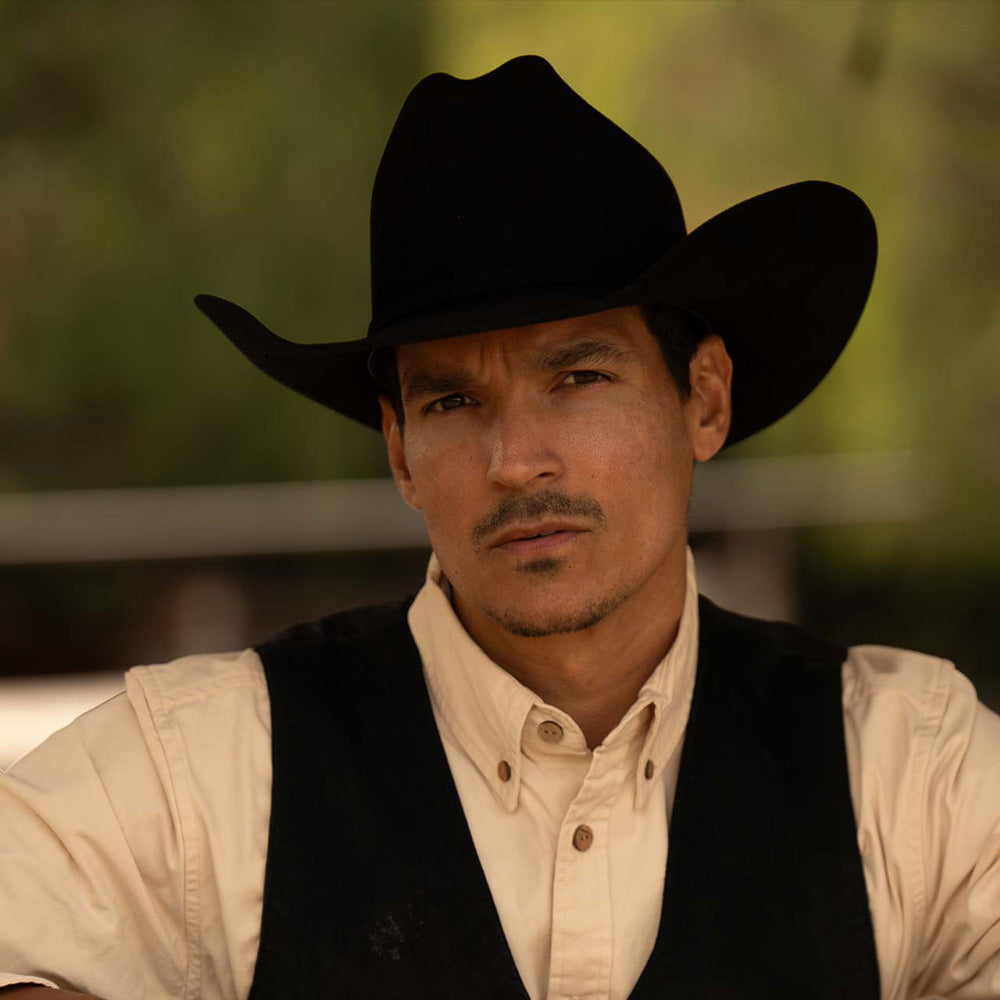 Man wearing a 6X Lockhart black cowboy hat with a black vest and white under shirt against a dark green background