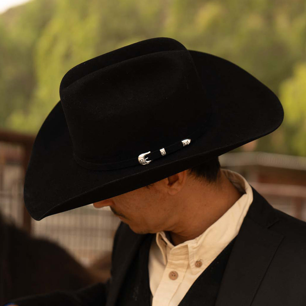 Man wearing a 6X Lockhart black cowboy hat with a black jacket and white under shirt against a dark green background while turning his head to the left to show the silver buckle on the band