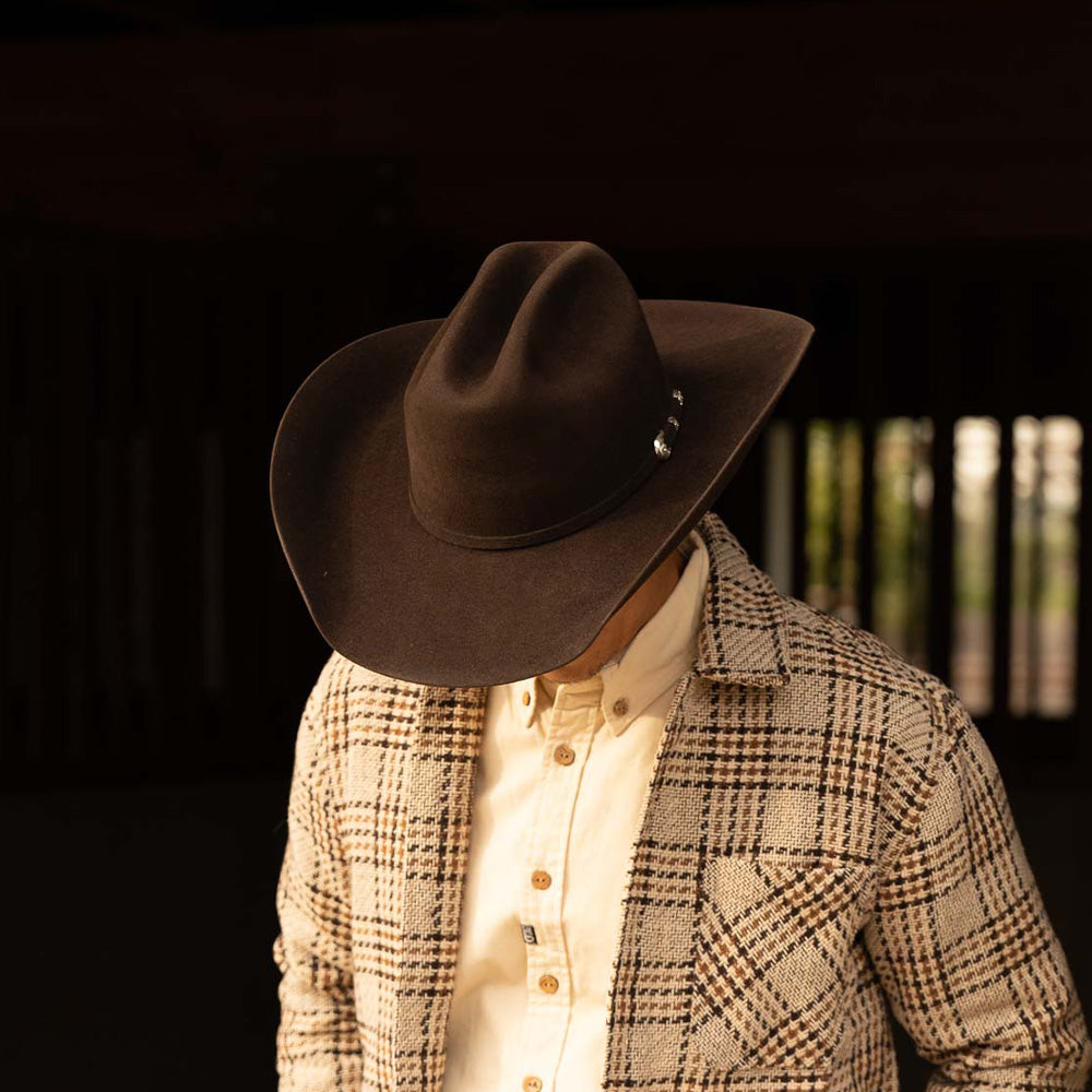 Man wearing a 6X Lockhart brown cowboy hat with a tan plaid jacket and white under shirt looking downwards to show the silver buckle on the band