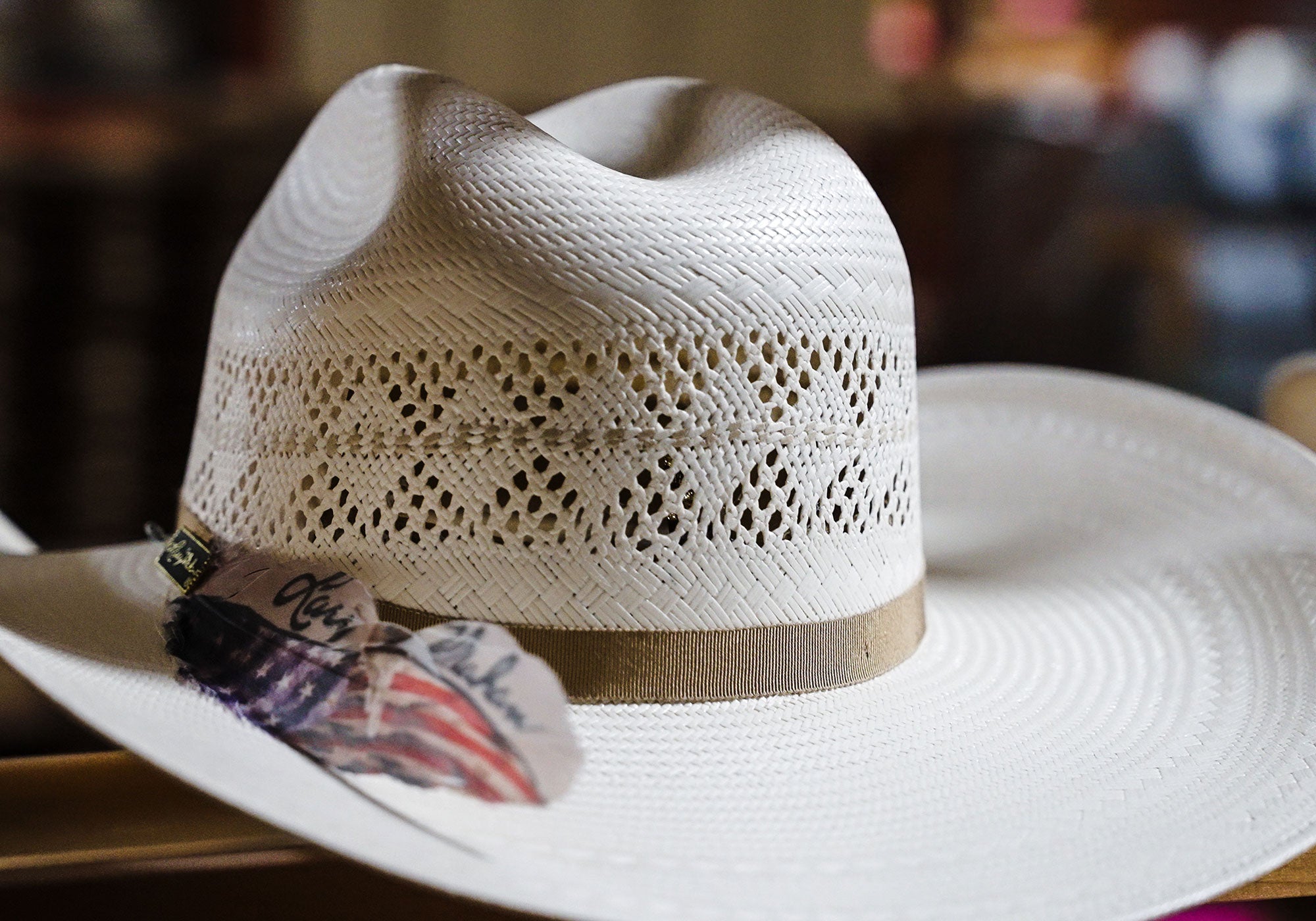 White Larry Mahan cowboy hat with a brown band and feather with the American flag printed along the side of it with the Larry Mahan logo in black