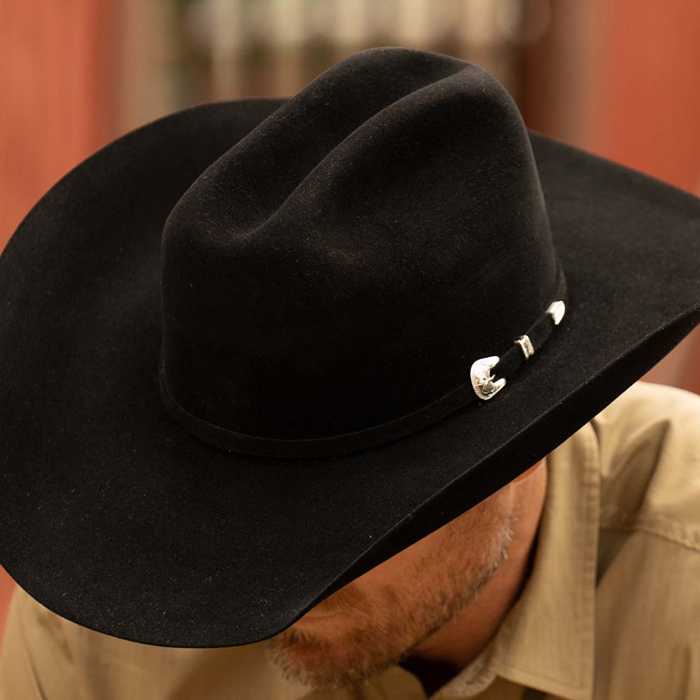 Bearded man wearing a 20X Pinnacle black cowboy hat and tan shirt standing in front of a red and brown barn looking down and to the left