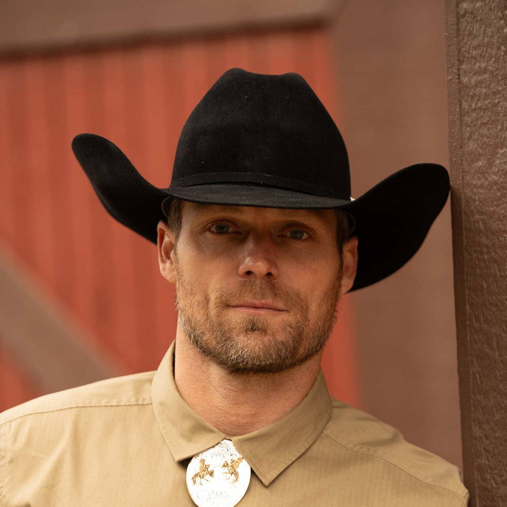 Bearded man wearing a 20X Pinnacle black cowboy hat and tan shirt with a shiny metalilc and gold bolo tie standing in front of a red and brown barn