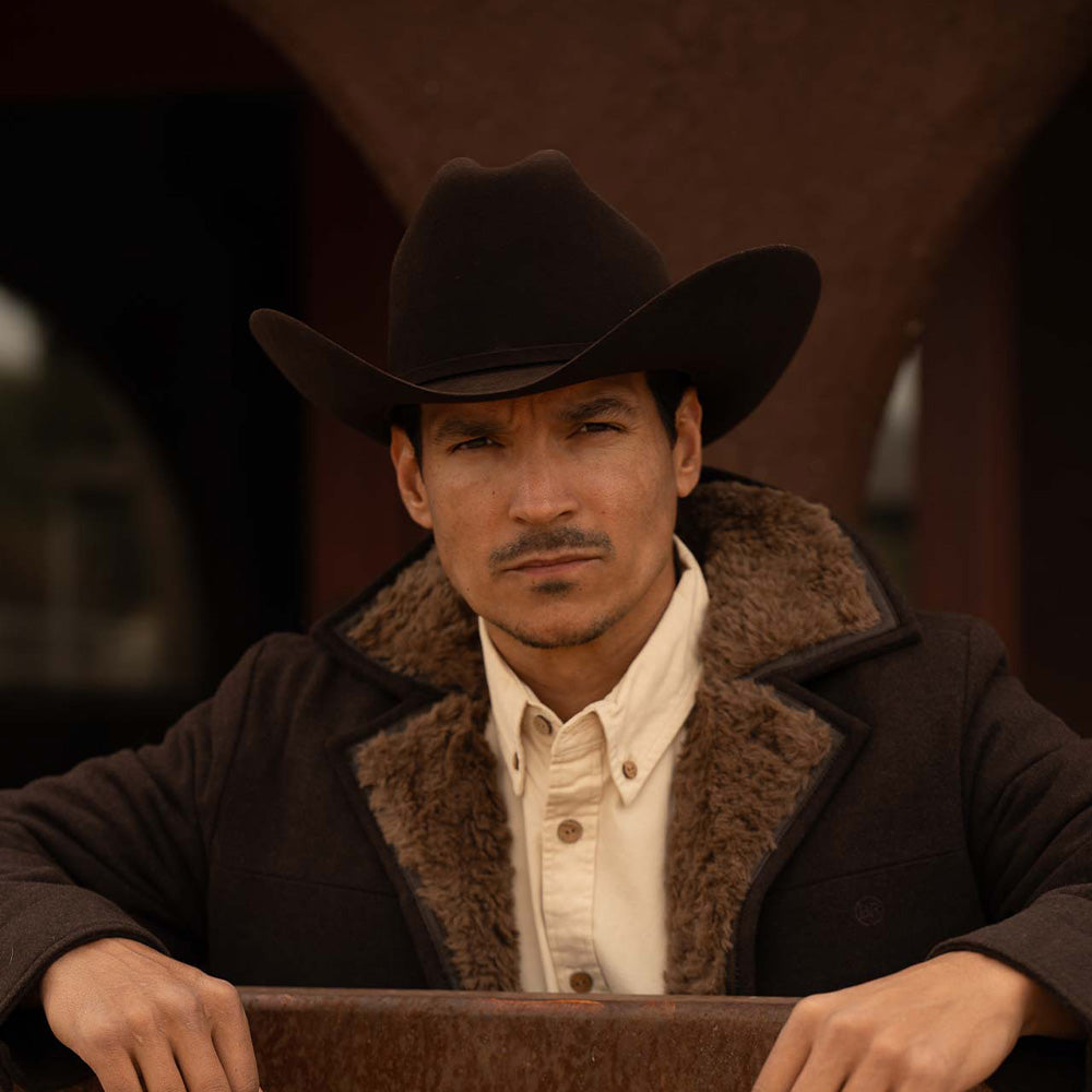 Man wearing a 6X Real brown cowboy hat, jacket with brown fur lining on the inside edges, and off-white shirt with both wrists resting on a metal beam