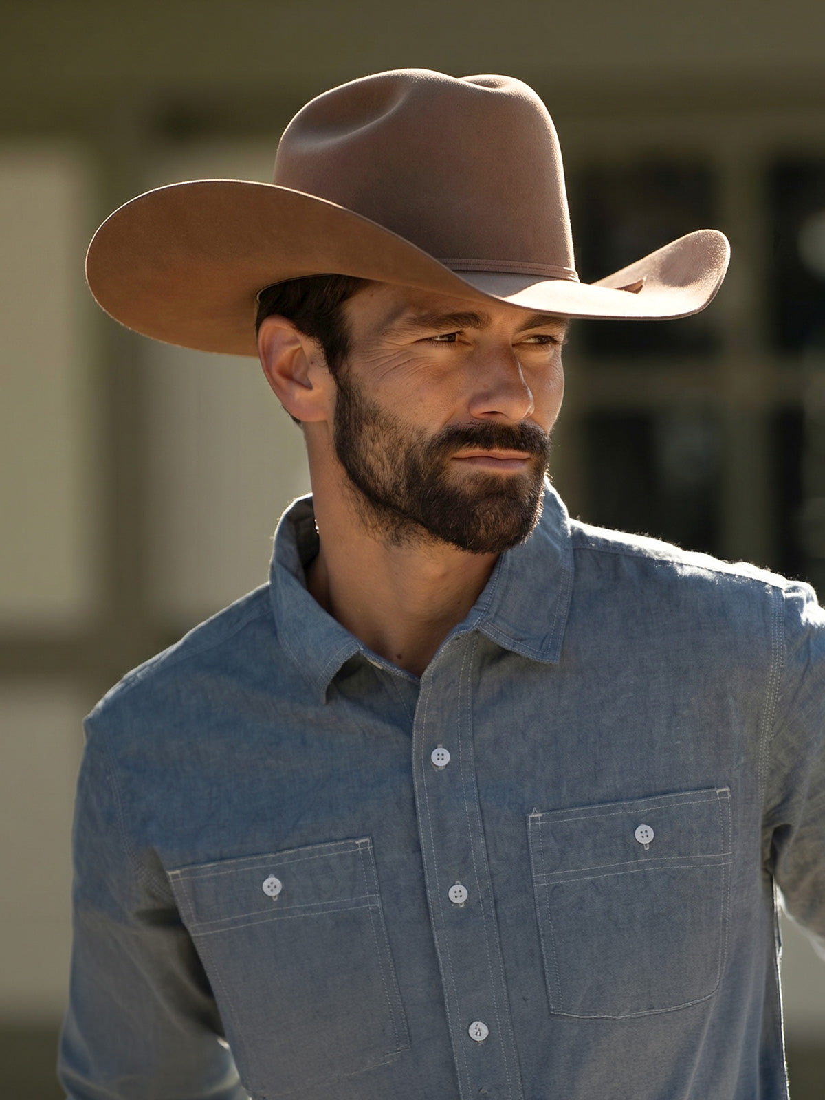 Bearded man wearing a tan colored cowboy hat and blue button up shirt standing near a blurred building wall in the background