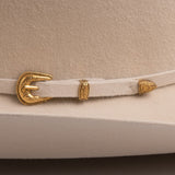 A close-up of a light beige felt hat band with ornate gold-tone metal hardware, including a decorative buckle and two matching keeper rings, displayed on a matching beige felt surface