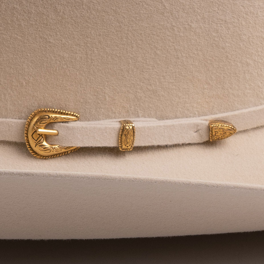A close-up of a light beige felt hat band with ornate gold-tone metal hardware, including a decorative buckle and two matching keeper rings, displayed on a matching beige felt surface