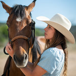 A woman in a belly colored cowboy hat and light blue top gently holds a brown horse outdoors under a clear blue sky.