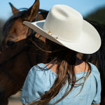 A woman seen from behind wearing a belly colored cowboy hat with a small gold accent and a light blue top, standing outdoors next to a brown horse.