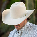 A man wearing a belly colored cowboy hat with a small gold decorative accent on the band, paired with a white patterned button-down shirt, set against a blurred outdoor background.