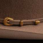 Close-up of a fawn brown felt hat with a matching brown strap and ornate gold-tone buckle and decorative accents
