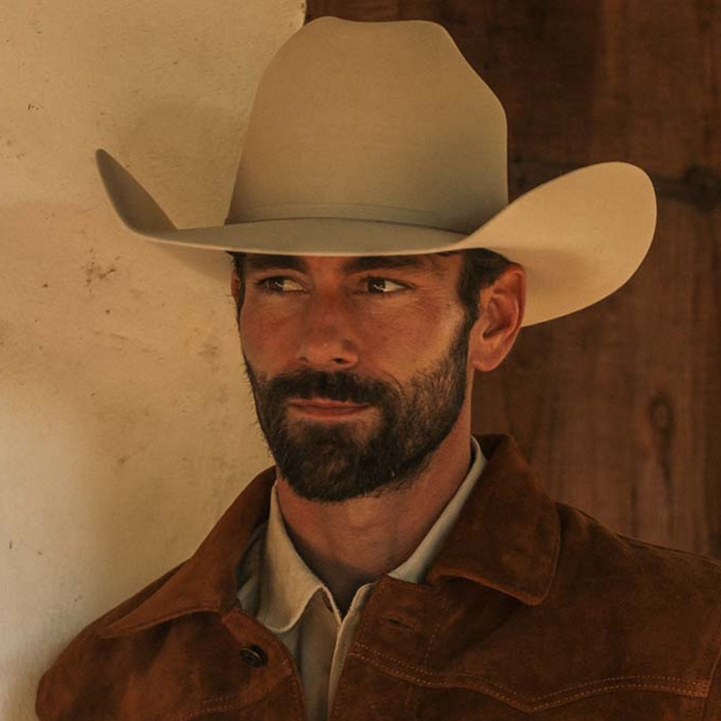 A man wearing a fawn colored cowboy hat and a brown leather jacket, looking off to the side against a rustic wall background.
