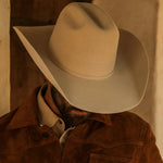 A close-up of a man wearing a fawn colored cowboy hat and a brown suede jacket, with his face obscured by the brim of the hat.