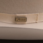 Close-up of a cream-colored felt hat with a matching grosgrain ribbon band and a central metal brand pin that reads "Worn by Legends"