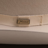 Close-up of a cream-colored felt hat with a matching grosgrain ribbon band and a central metal brand pin that reads "Worn by Legends"