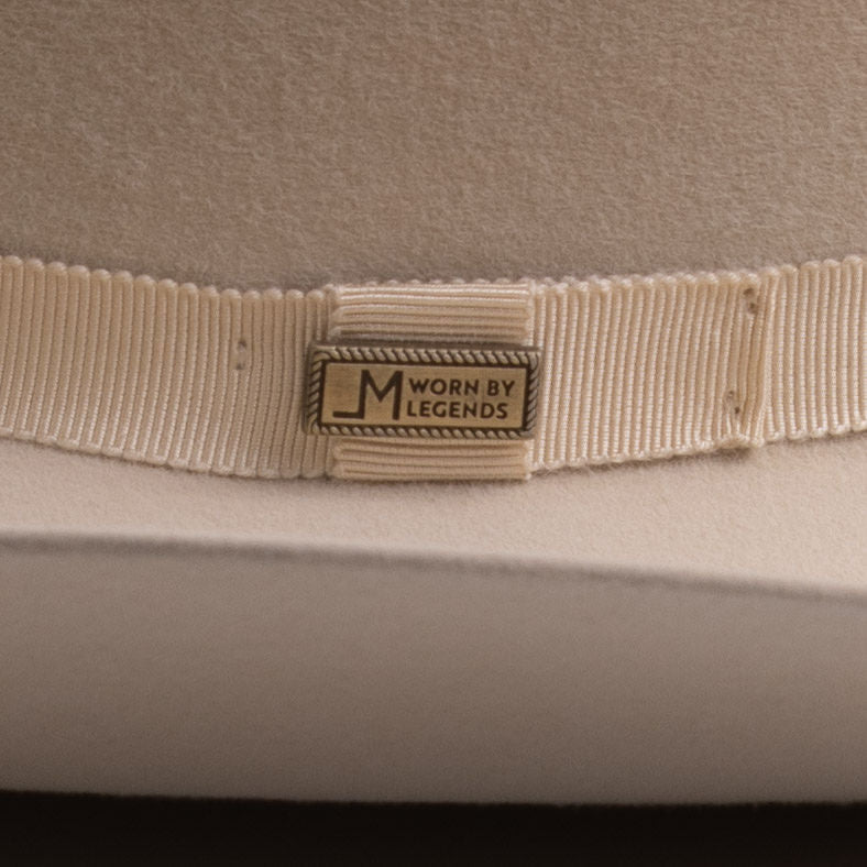 Close-up of a cream-colored felt hat with a matching grosgrain ribbon band and a central metal brand pin that reads "Worn by Legends"