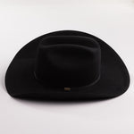 A black felt cowboy hat with a curved brim and a small gold brand plaque on the band, photographed on a light gray background