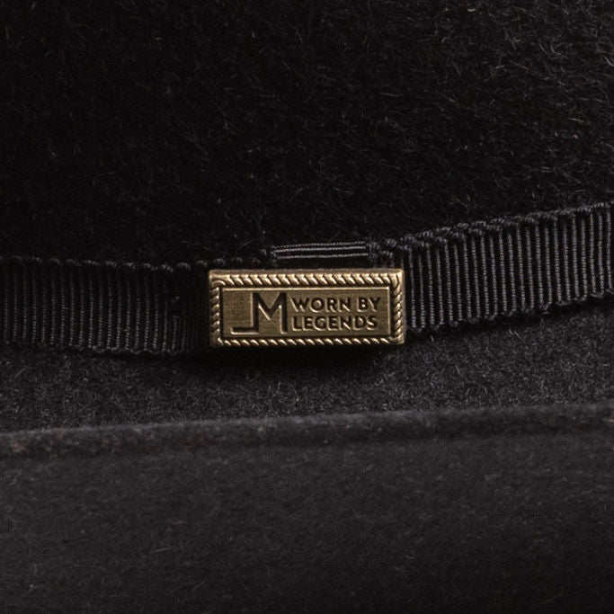 Close-up of a black felt hat with a black ribbon band and a rectangular gold-tone metal pin engraved with the brand "Worn by Legends"