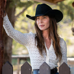 A woman wearing a black cowboy hat and a white textured knit long-sleeve top leans against a wooden post in an outdoor setting with trees and a fence visible.