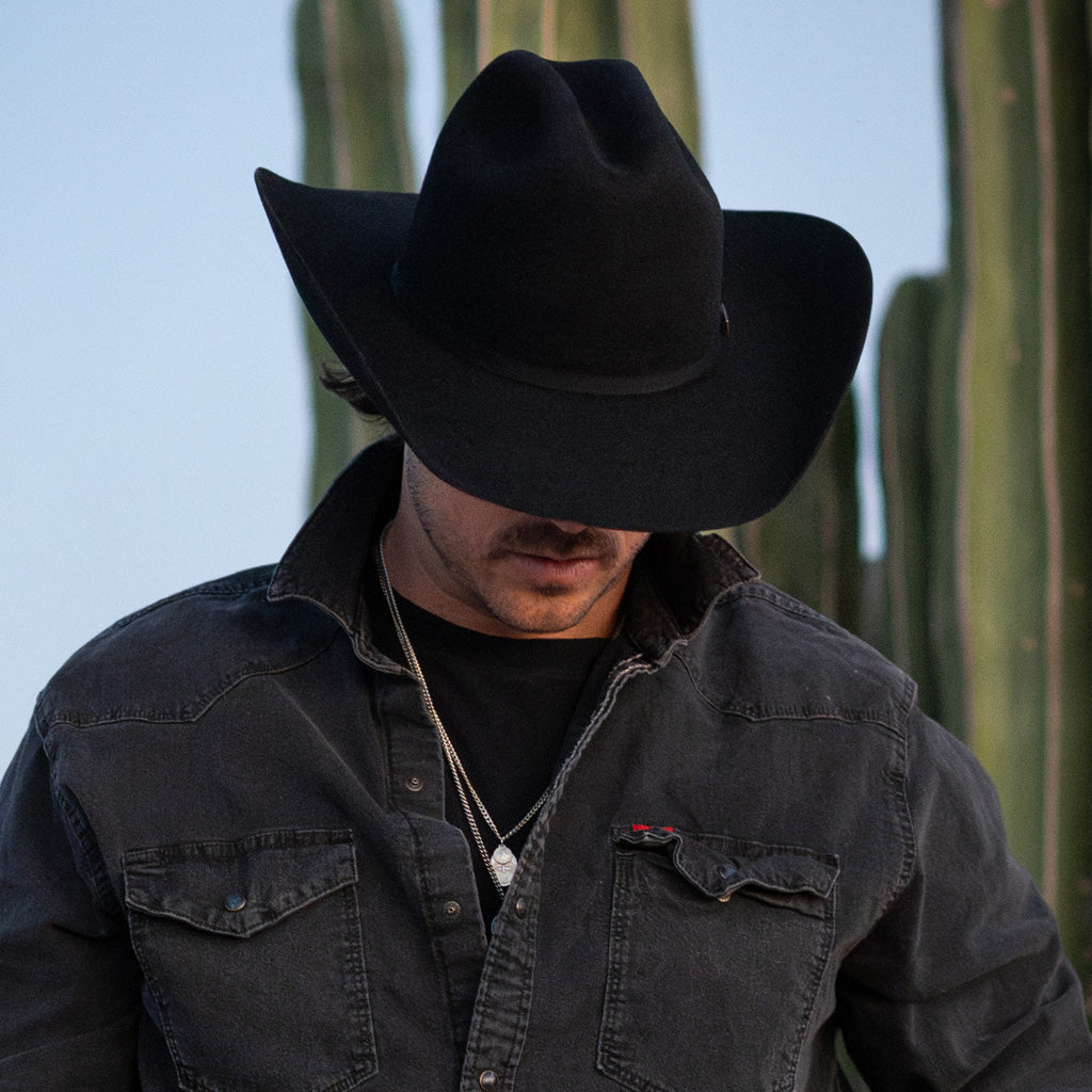 A man in a black cowboy hat and dark jacket outdoors leaning near a cactus.
