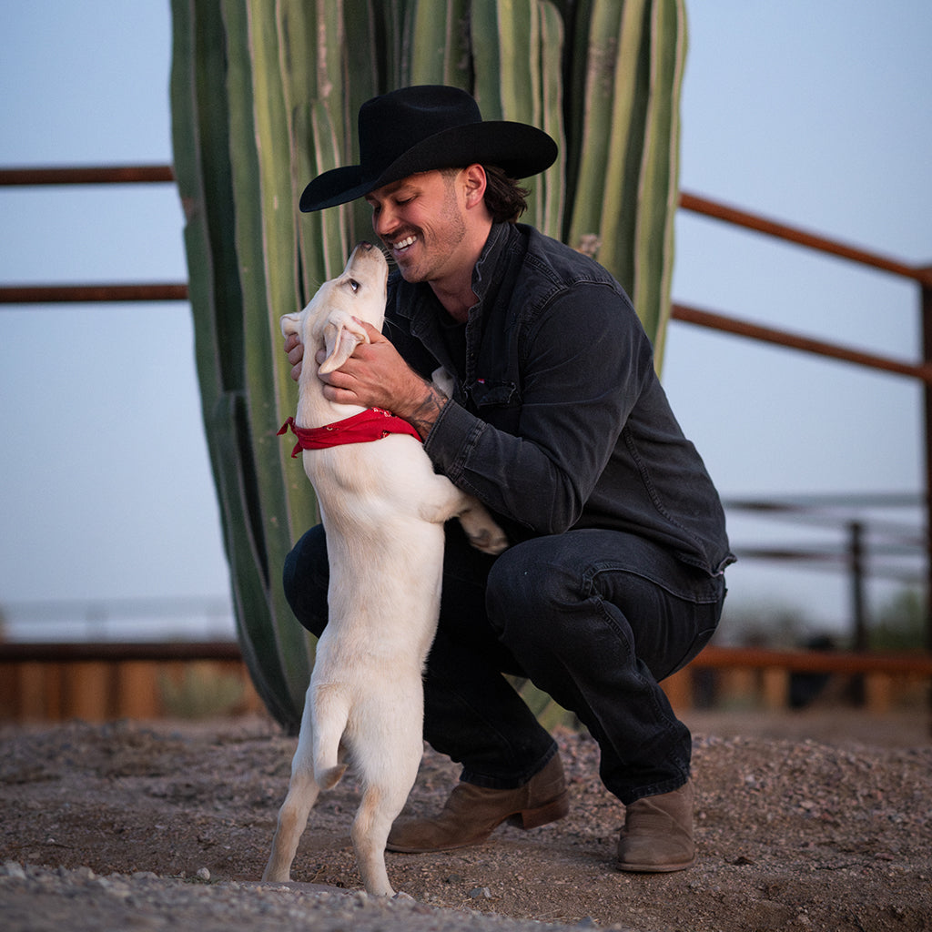 A smiling man in a black cowboy hat and dark shirt crouches on a dirt ground next to a large saguaro cactus, hugging a white dog that is standing on its hind legs and wearing a red bandana.
