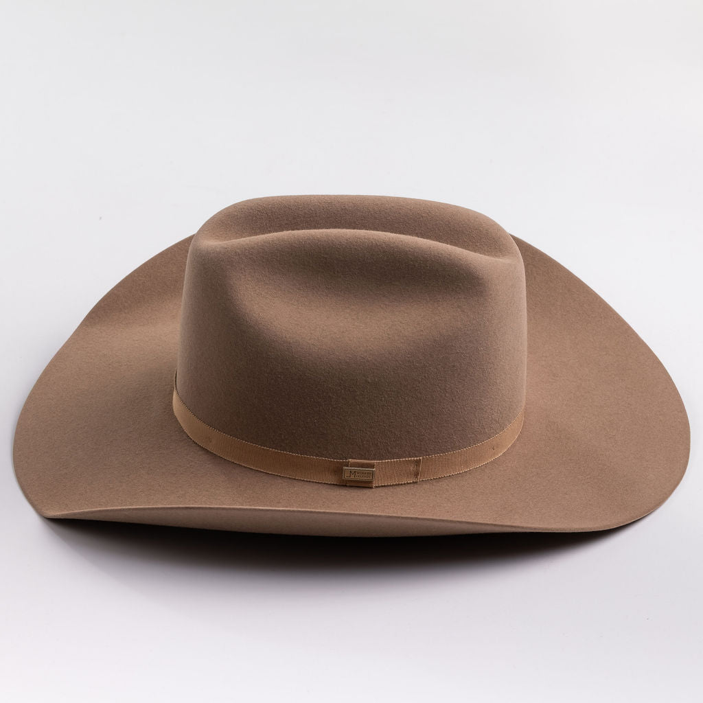 A classic felt cowboy hat in a fawn brown color, featuring a matching band with a small gold-tone metal buckle, photographed on a light gray background