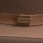 Close-up of a brown felt hat with a matching brown grosgrain ribbon band and a brass-tone metal badge engraved with "LM WORN BY LEGENDS"