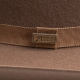Close-up of a brown felt hat with a matching brown grosgrain ribbon band and a brass-tone metal badge engraved with "LM WORN BY LEGENDS"