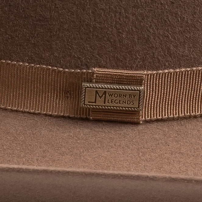 Close-up of a brown felt hat with a matching brown grosgrain ribbon band and a brass-tone metal badge engraved with "LM WORN BY LEGENDS"