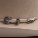 Close-up of a light beige felt cowboy hat featuring a decorative silver-toned metal buckle and strap closure