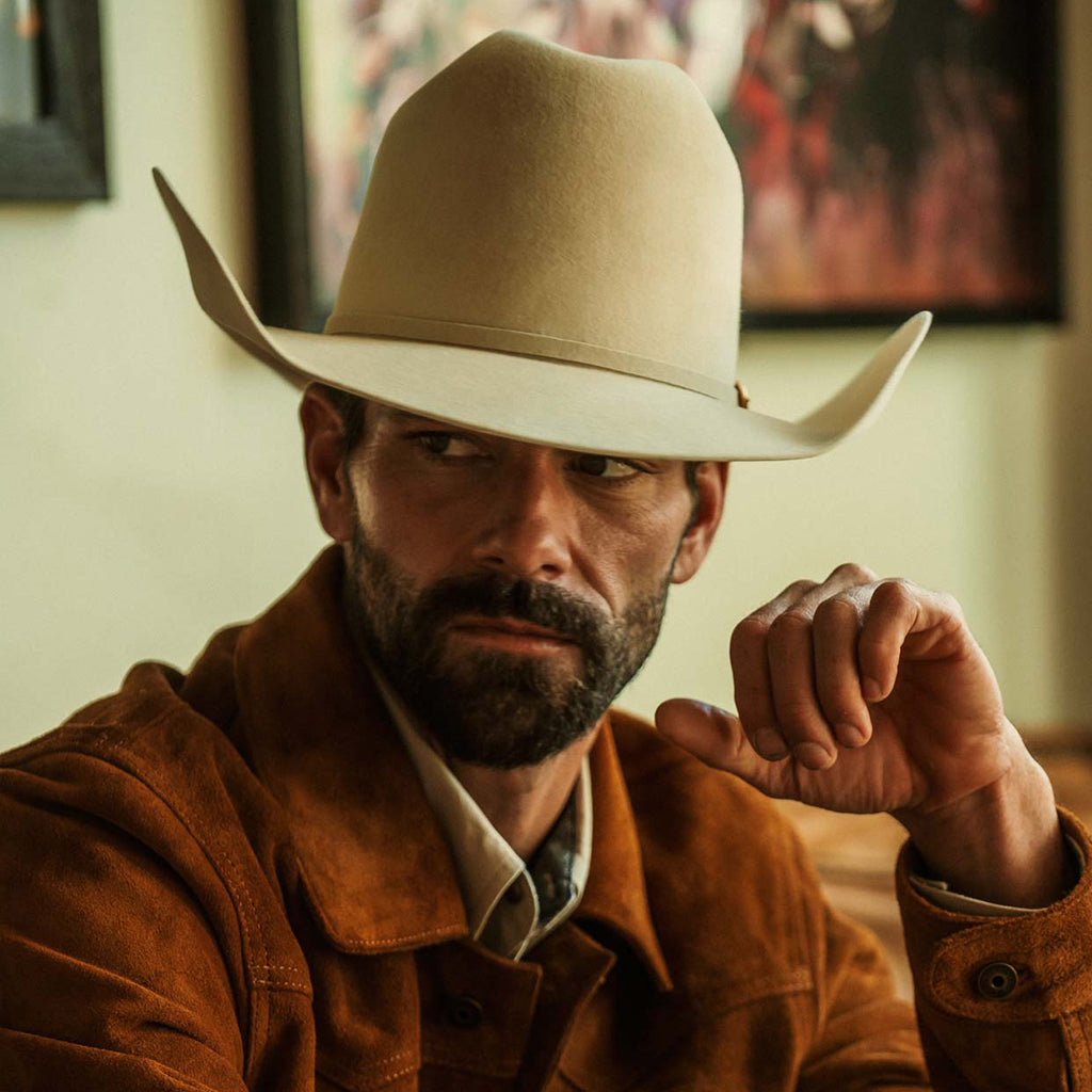 A close-up portrait of a man wearing a belly colored cowboy hat and a brown suede jacket, with a blurred indoor background and framed wall art.