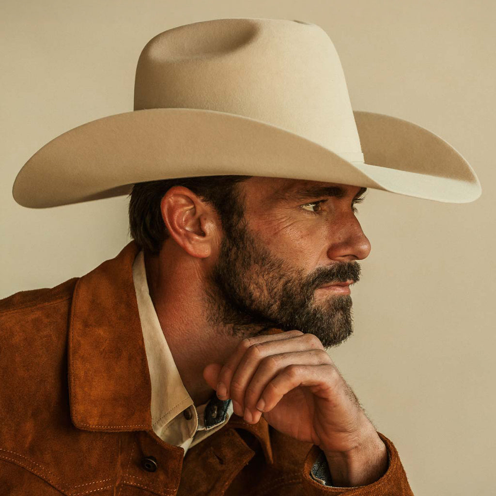 A close-up portrait of a man wearing a belly colored cowboy hat and a brown suede jacket over a cream shirt, posed against a solid beige background.