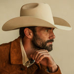 A close-up portrait of a man wearing a belly colored cowboy hat and a brown suede jacket over a cream shirt, posed against a solid beige background.