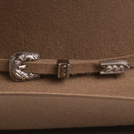 A close-up of a brown felt hat featuring a decorative silver-toned concho buckle and matching silver-toned tip hardware on the strap