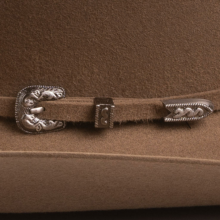A close-up of a brown felt hat featuring a decorative silver-toned concho buckle and matching silver-toned tip hardware on the strap