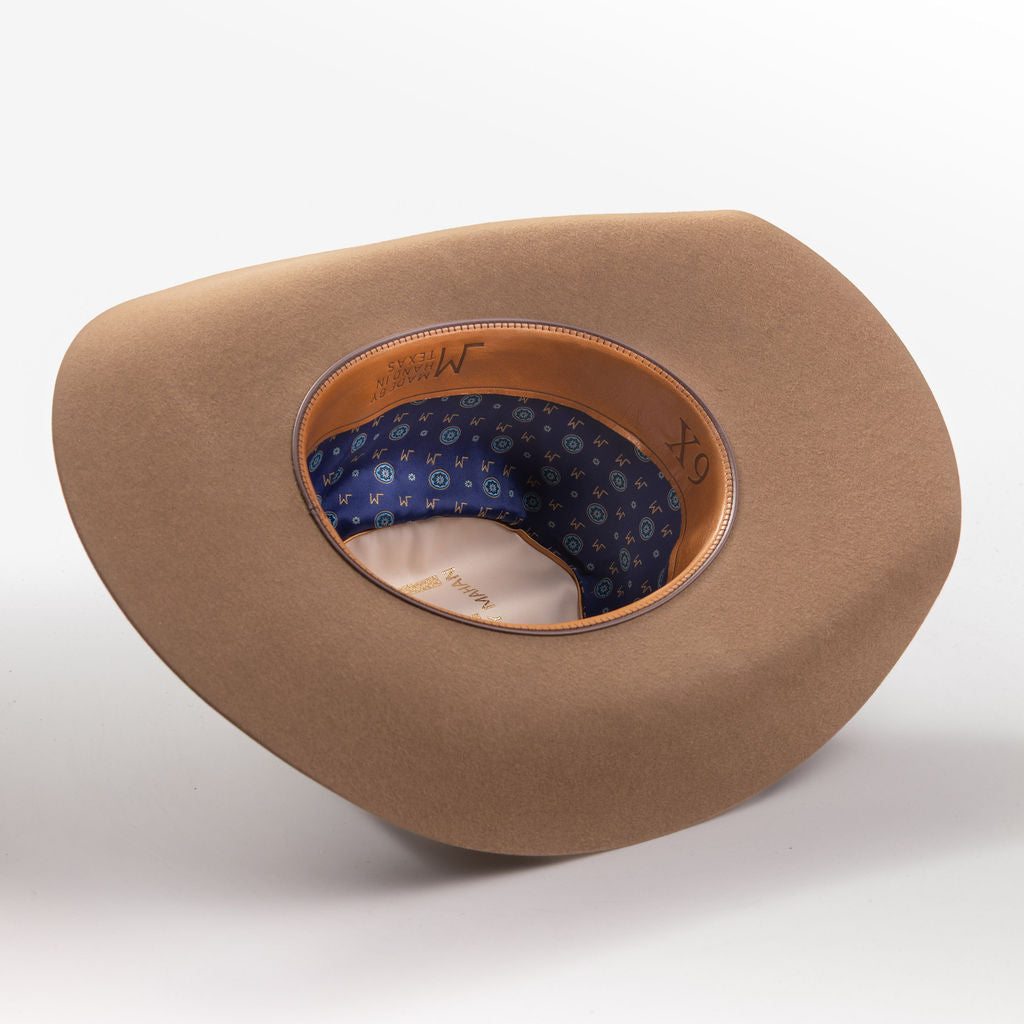A tan cowboy hat with a wide brim, shown from a three-quarters perspective to reveal the interior; the hat features a patterned blue inner band with small white stars and a tan leather trim