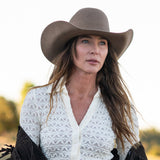 A woman wearing a fawn colored cowboy hat and a white textured knit top, photographed outdoors against a bright, softly blurred background.