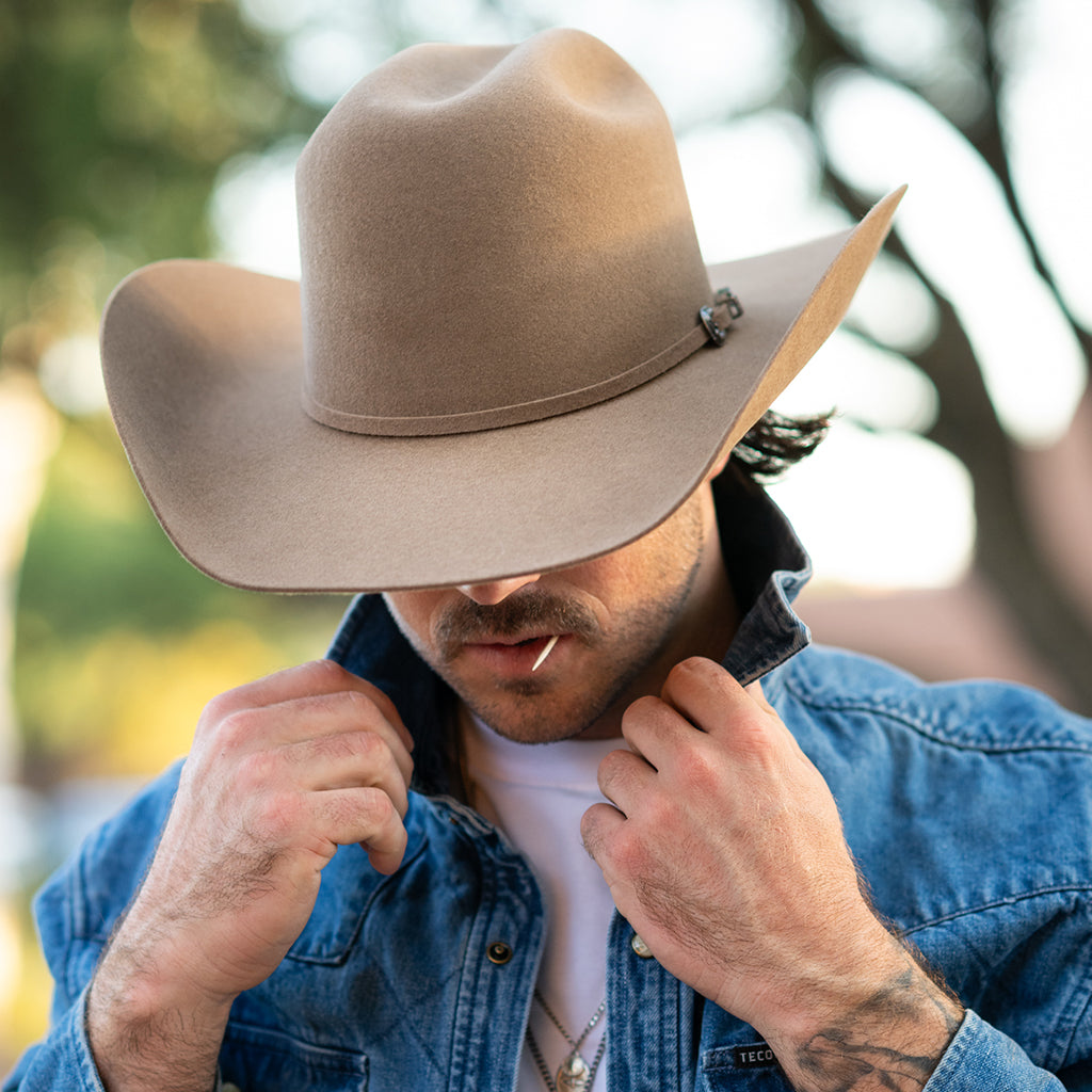 A man wearing a fawn colored felt cowboy hat with a small decorative pin and a blue denim jacket, adjusting the collar of his shirt in an outdoor setting.